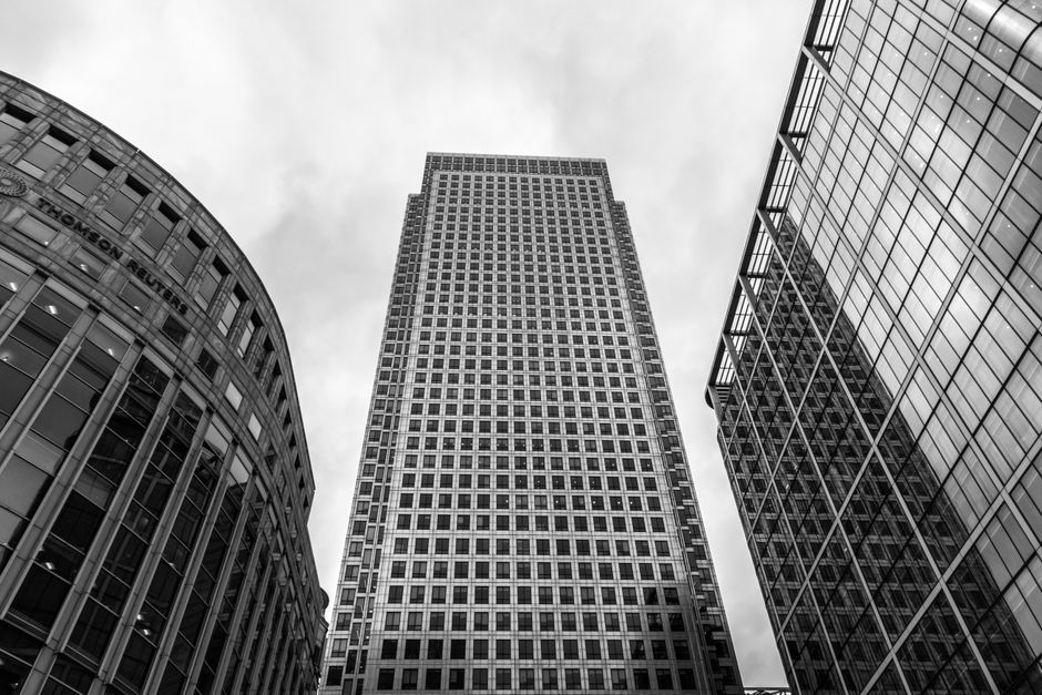 A black and white photograph taken from a low angle showing three modern office buildings in Canary Wharf. The central building is a tall rectangular skyscraper with a grid-like pattern of windows covering its facade, surrounded by two shorter, rounded buildings with curved glass exteriors and visible window frames. In the foreground, visible inside a property, are several cardboard boxes stacked near a doorway. Behind the boxes, a moving blanket is draped over an item to protect it during transport. The space appears to be a hallway or an entryway with a view of the exterior through an open door or window. The image captures the process of home relocation or furniture transport, emphasizing the steps involved in packing and preparing items for moving, associated with professional removals services provided by Man with Van Canary Wharf.