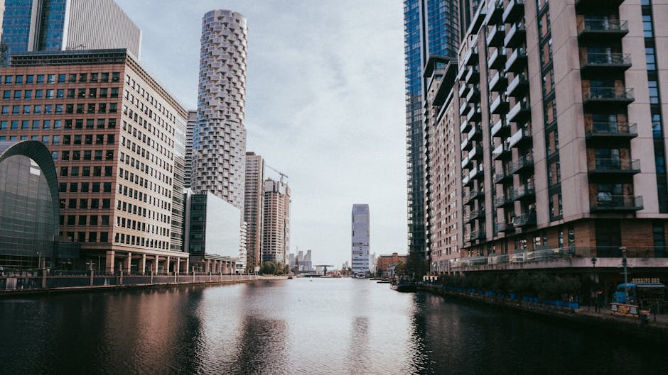 A cityscape view along Canary Wharf featuring tall modern office buildings with a mix of glass and concrete facades, situated beside a calm waterway with reflections of the buildings on the surface. The sky is partly cloudy with soft natural light illuminating the scene. Behind the water, the harbor area includes a loading zone where a black van is parked, partially visible to the right. Inside the van, visible tools such as moving blankets, cardboard boxes, and packing materials suggest an ongoing home relocation or furniture transport process. A person wearing casual clothing is seen lifting a cardboard box onto the vehicle's loading area, assisted by a trolley or hand truck. The scene captures elements typical of house removals in the area, with the urban environment highlighting the logistics involved in packing, loading, and transporting belongings using professional services like those provided by Man with Van Canary Wharf, situated in a modern city setting.