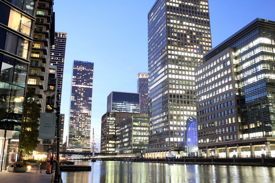 A black and white photograph taken from a low angle showing three modern office buildings in Canary Wharf. The central building is a tall rectangular skyscraper with a grid-like pattern of windows covering its facade, surrounded by two shorter, rounded buildings with curved glass exteriors and visible window frames. In the foreground, visible inside a property, are several cardboard boxes stacked near a doorway. Behind the boxes, a moving blanket is draped over an item to protect it during transport. The space appears to be a hallway or an entryway with a view of the exterior through an open door or window. The image captures the process of home relocation or furniture transport, emphasizing the steps involved in packing and preparing items for moving, associated with professional removals services provided by Man with Van Canary Wharf.