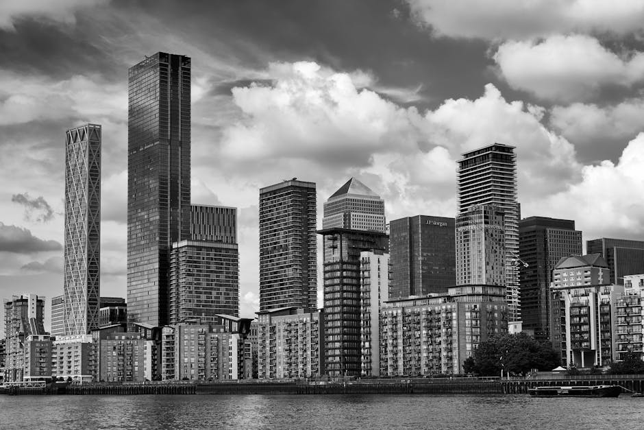 A black and white photograph of a modern city skyline with tall skyscrapers and high-rise buildings, positioned along a waterfront. The buildings feature various architectural styles, including glass facades, grid-patterned exteriors, and geometric designs. Some structures are under construction with scaffolding and cranes visible. The skyline is set against a partly cloudy sky with substantial cloud cover. In the foreground, a promenade or quay runs parallel to the water, with a few trees and a small dock visible. This scene captures the urban environment typical for house removals or furniture transport as part of a home relocation process, illustrating the dense cluster of buildings that residents and moving services in Canary Wharf might navigate. Occasionally, a moving company such as Man with Van Canary Wharf might be involved in this type of urban logistics to facilitate packing, loading, and transporting belongings through such cityscapes.