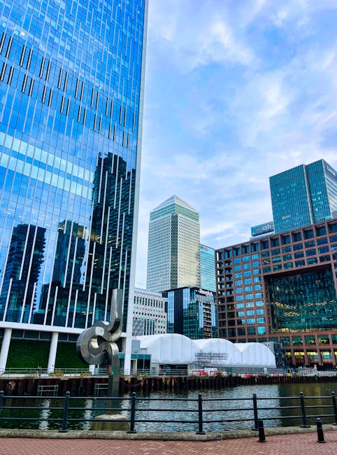 A daytime view of Canary Wharf in London showing a collection of tall, modern office buildings with glass facades reflecting the sky and surrounding structures. In the foreground, there is a waterway lined with a black metal railing, with a metallic sculpture situated near the edge. Inside the property, visible through an open doorway, are cardboard boxes sealed with brown packing tape stacked on a wooden platform, along with rolls of bubble wrap and several plastic containers used for packing and protecting furniture during home relocation. The lighting is natural, highlighting the intricate details of the buildings and the packing materials, as Man with Van Canary Wharf prepares for furniture transport or moving services. The overall scene emphasizes the logistical process involved in packing and loading belongings for efficient house removals within the Canary Wharf area.