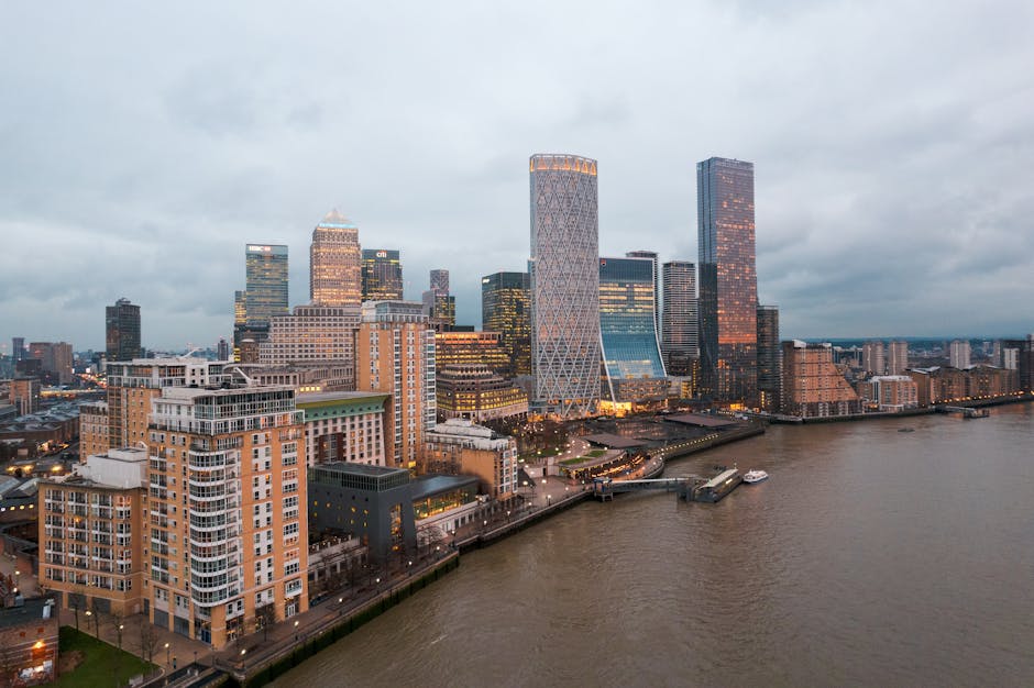 A black and white photograph of a modern city skyline with tall skyscrapers and high-rise buildings, positioned along a waterfront. The buildings feature various architectural styles, including glass facades, grid-patterned exteriors, and geometric designs. Some structures are under construction with scaffolding and cranes visible. The skyline is set against a partly cloudy sky with substantial cloud cover. In the foreground, a promenade or quay runs parallel to the water, with a few trees and a small dock visible. This scene captures the urban environment typical for house removals or furniture transport as part of a home relocation process, illustrating the dense cluster of buildings that residents and moving services in Canary Wharf might navigate. Occasionally, a moving company such as Man with Van Canary Wharf might be involved in this type of urban logistics to facilitate packing, loading, and transporting belongings through such cityscapes.
