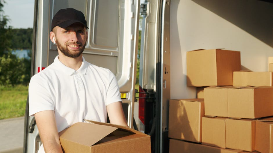 A man dressed in a blue beanie, navy and red jacket, black pants, and white sneakers is sitting on the edge of an open van during a house relocation process. Behind him, the van's interior is visible,